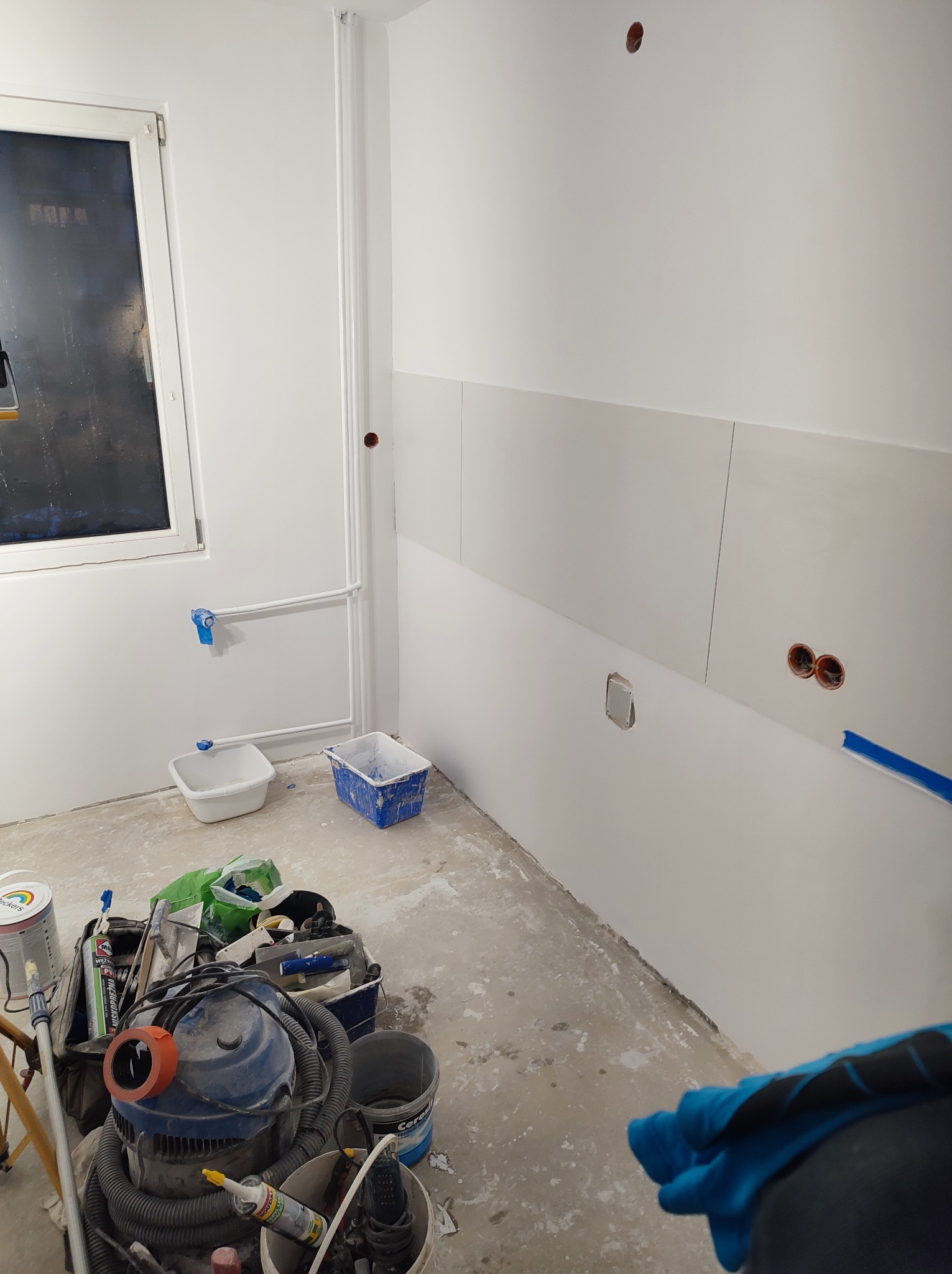 Wnętrze w trakcie prac w Tarnobrzegu: białe ściany, częściowo wykończone płyty, zabezpieczone rury, widoczny odkurzacz przemysłowy i narzędzia malarskie na podłodze.