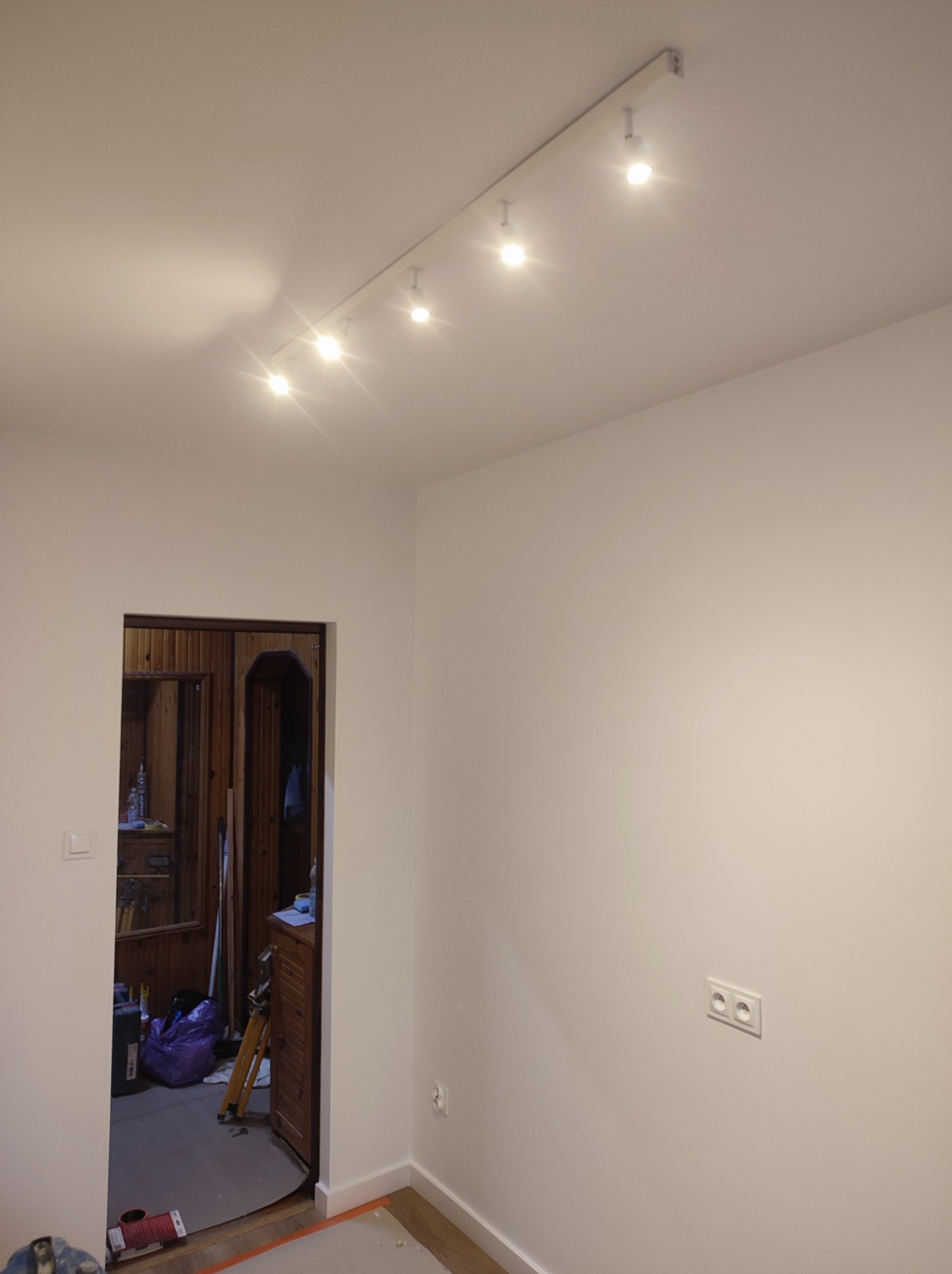 Białe ściany po malowaniu z widoczną listwą przypodłogową i oświetleniem sufitowym na szynie, w tle widoczne otwarte drzwi do zagraconego pokoju z boazerią.