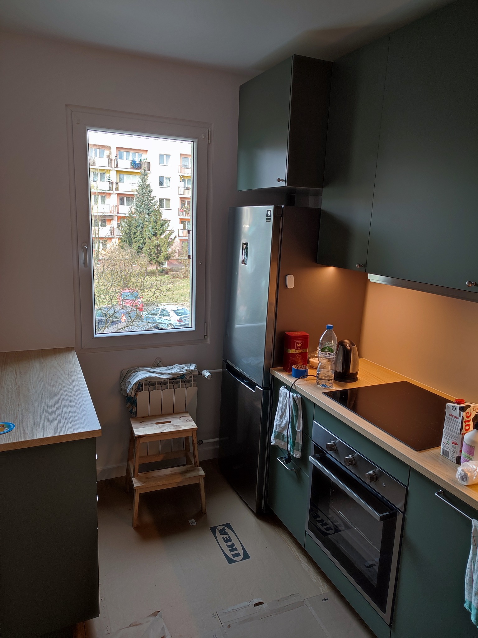 Nowoczesna kuchnia w odcieniach zieleni z drewnianymi blatami, widok na blok z okna, na podłodze kartony zabezpieczające podłogę, drabinka oparta o grzejnik pod oknem.