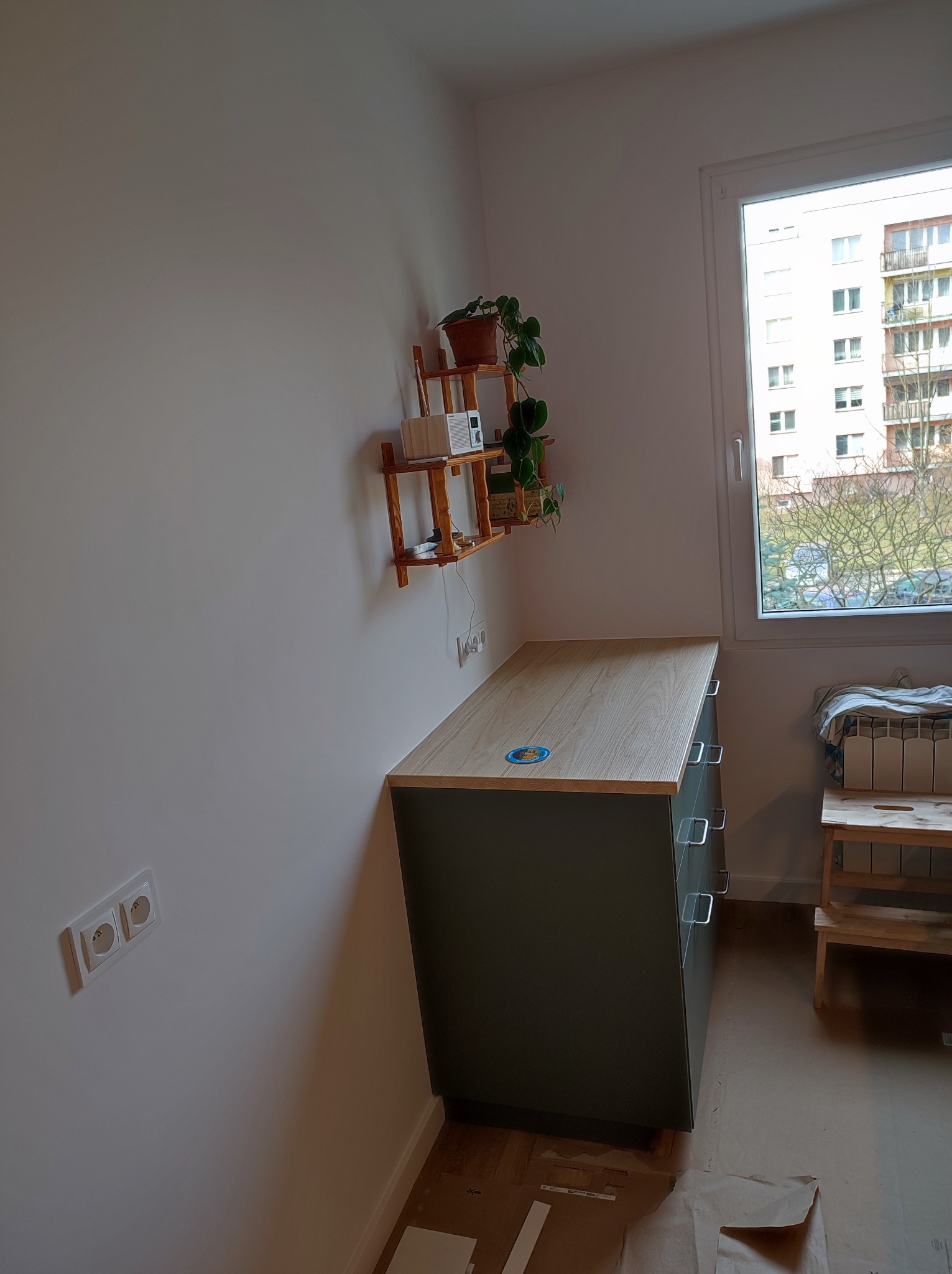 Szafka z szufladami o drewnianym blacie, stojąca pod ścianą z półką na kwiaty, widoczne gniazdko elektryczne, okno z widokiem na budynek mieszkalny i zabezpieczona podłoga.