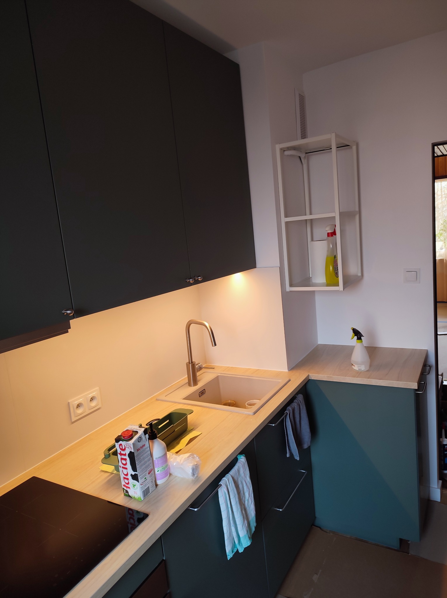 Nowoczesna kuchnia z ciemnozielonymi szafkami, drewnianym blatem, stalową baterią i widocznymi produktami spożywczymi na blacie. Oświetlenie podszafkowe dodaje ciepła, a biała półka na ścianie...