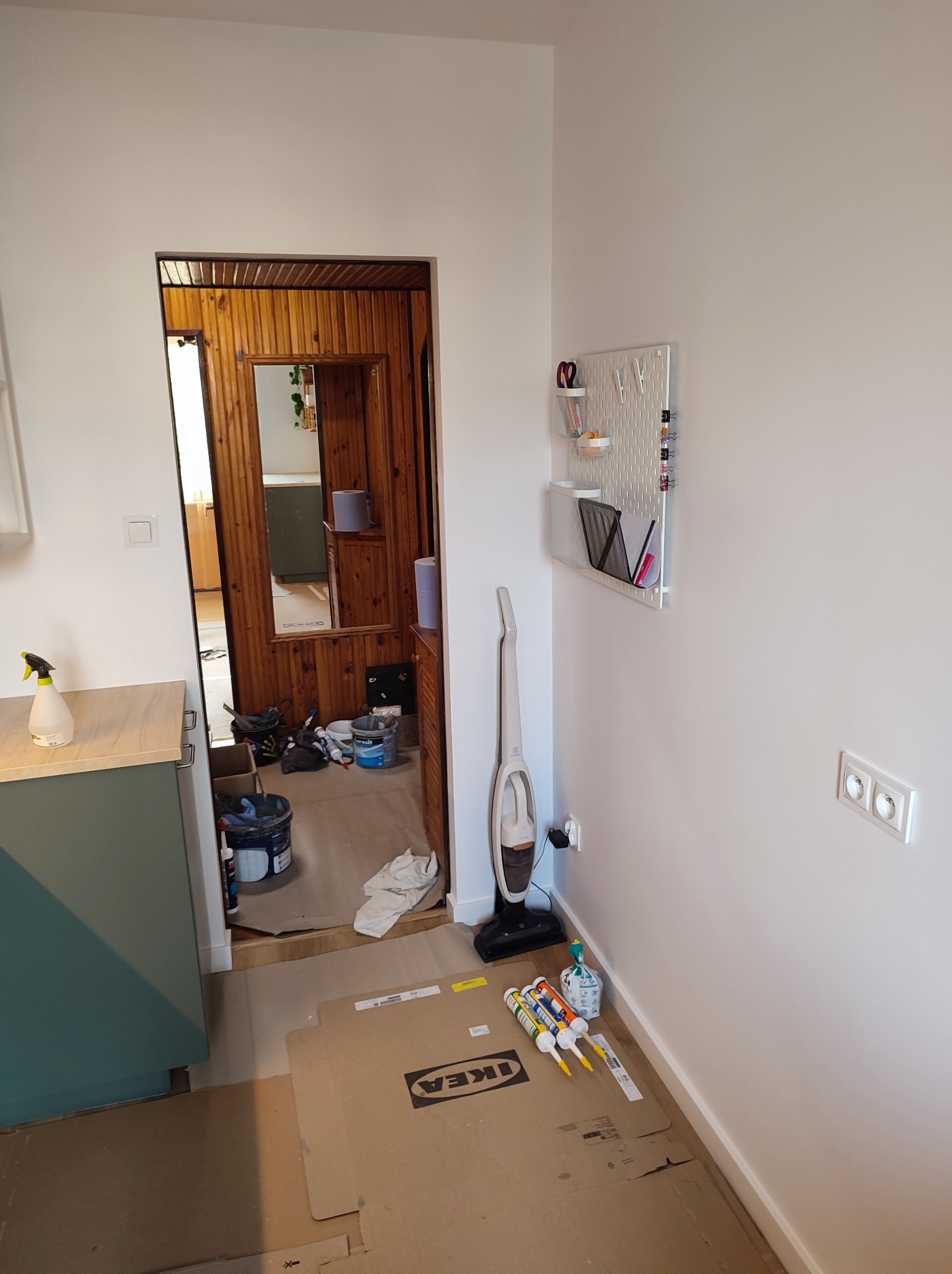 Wnętrze w trakcie prac: widoczne wiadro z farbą, pędzle, kartony ochronne IKEA, odkurzacz pionowy i listwa zasilająca na ścianie.