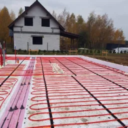 Fundamenty z ogrzewaniem podłogowym w domu jednorodzinnym w trakcie budowy. Widoczne czerwone rury, styropian i ściany budynku w tle.