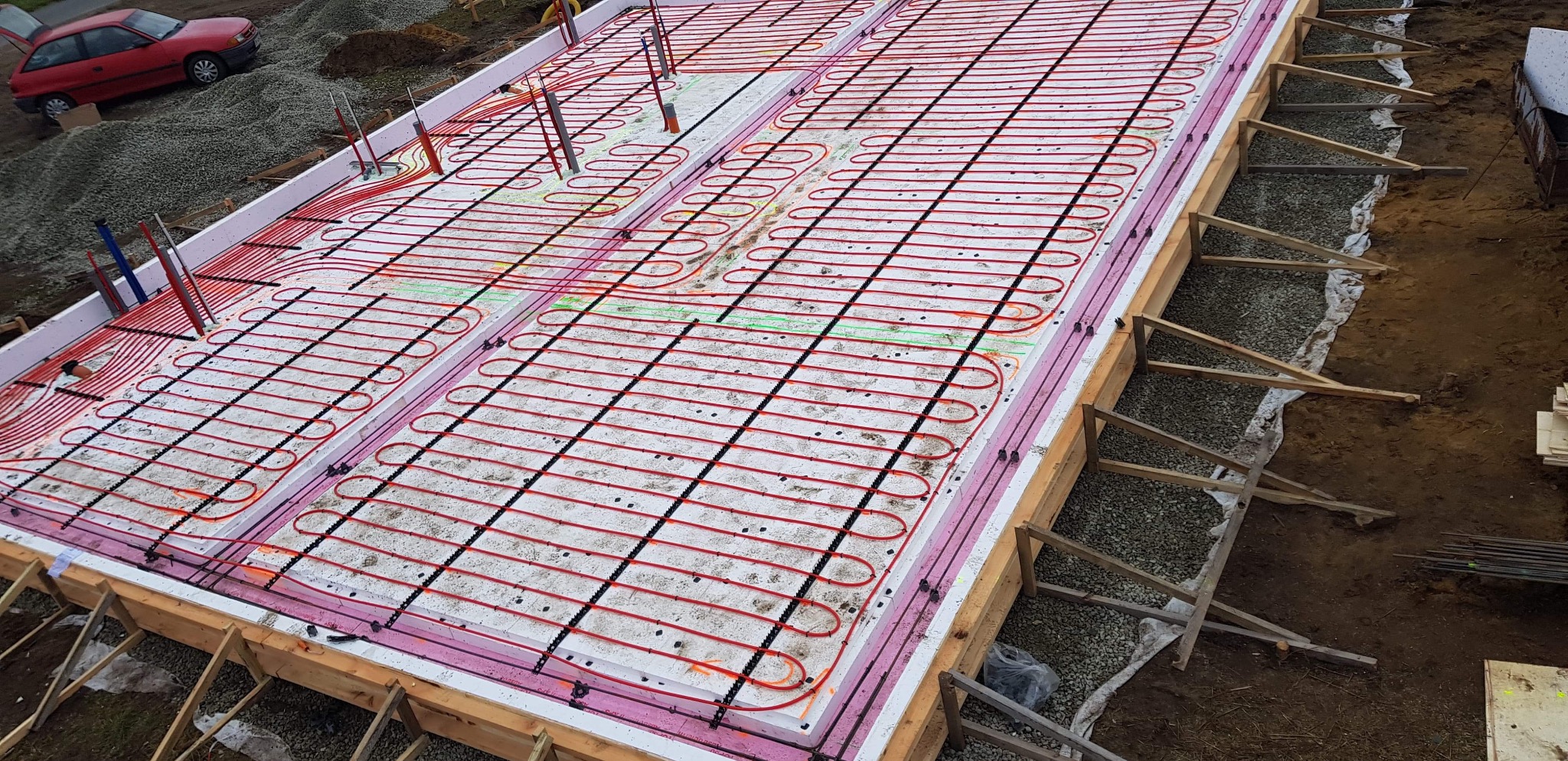 Fundamenty z izolacją i ogrzewaniem podłogowym w trakcie montażu. Widoczne czerwone rury grzewcze ułożone na białym styropianie, szalunek drewniany i podsypka żwirowa.