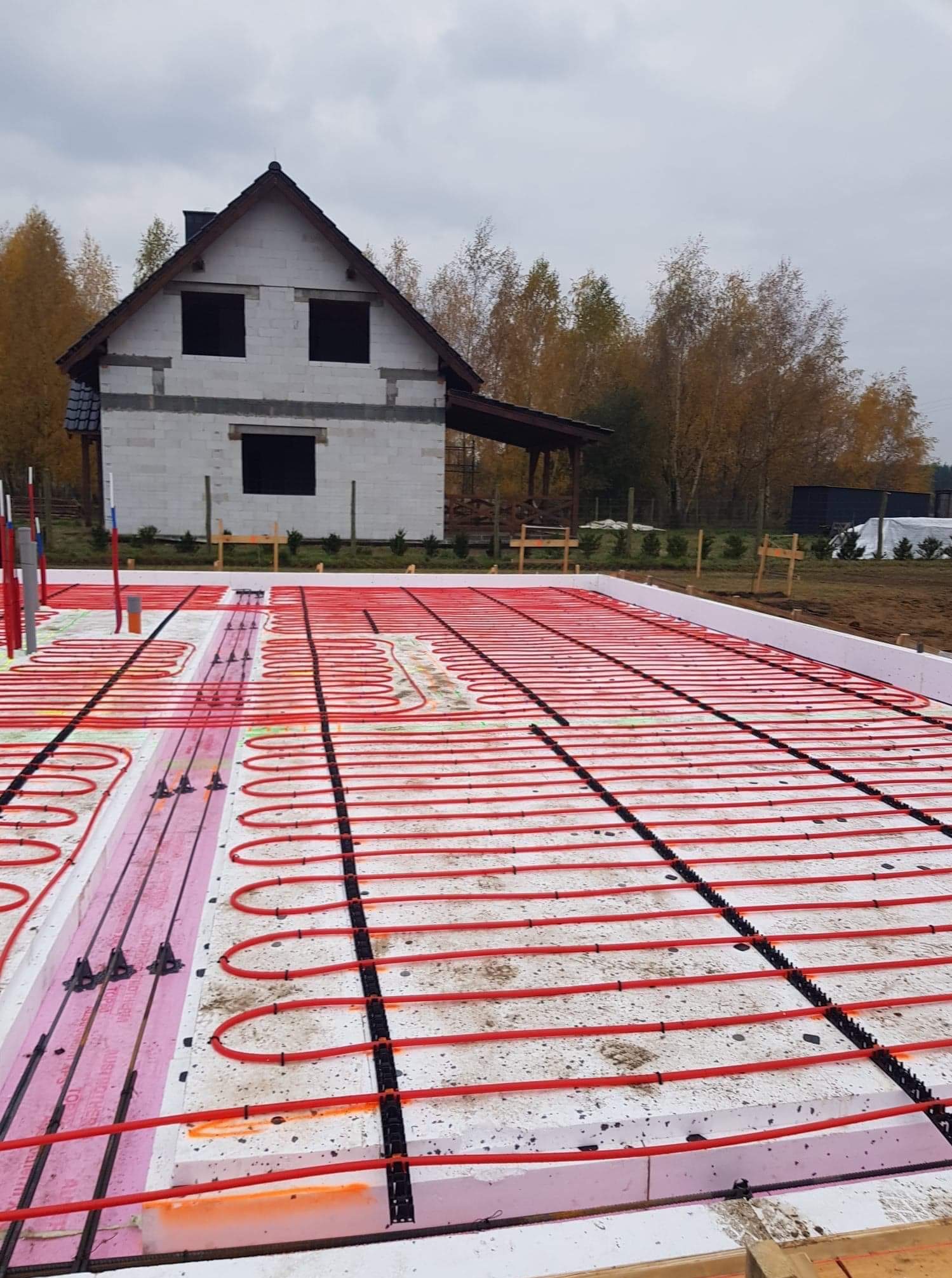 Fundamenty z ogrzewaniem podłogowym w domu jednorodzinnym w trakcie budowy. Widoczne czerwone rury, styropian i ściany budynku w tle.
