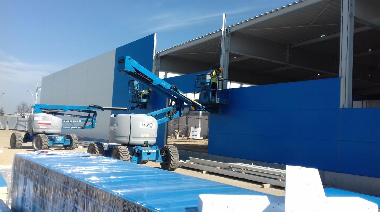 Montaż niebieskich paneli ściennych na konstrukcji hali przemysłowej z użyciem podnośników koszowych, widoczne stalowe elementy konstrukcyjne dachu.