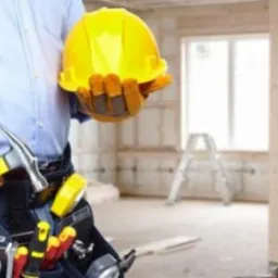 Mężczyzna w niebieskiej koszuli i rękawicach roboczych trzyma żółty kask i młotek, widoczny pas z narzędziami, w tle surowy stan wykończenia wnętrza z widocznymi schodami drabinkowymi.