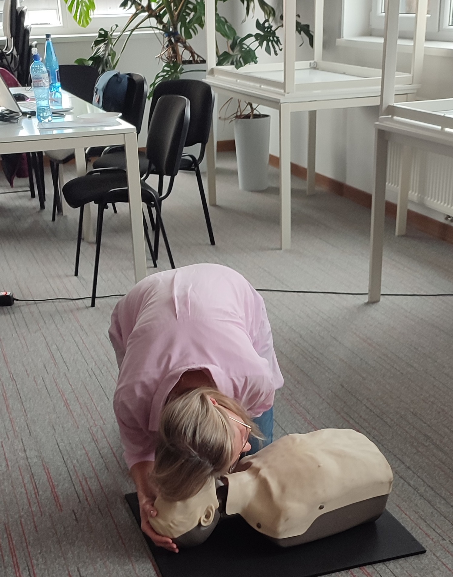 Kobieta w różowej koszuli demonstruje resuscytację na manekinie na podłodze w biurze, widoczne biurka, krzesła i rośliny doniczkowe w tle.