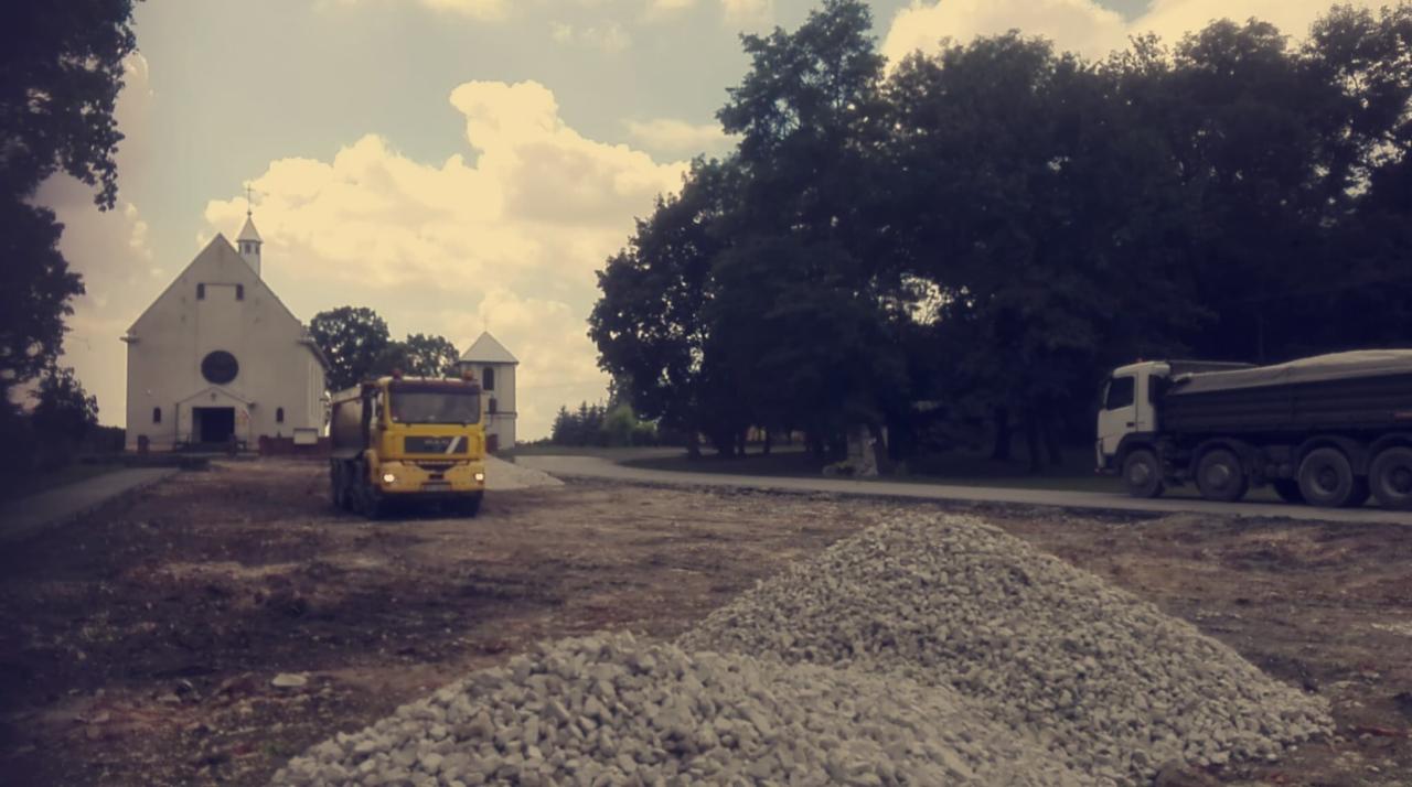 Żółta wywrotka Volvo na placu budowy przed białym kościołem, w tle druga wywrotka i hałda gruzu.