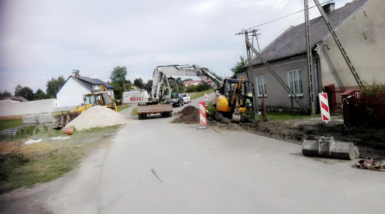 Dwie koparki pracują przy drodze w Pińczowie, widoczny wykop i sterta ziemi, w tle budynki mieszkalne i zaparkowane samochody.