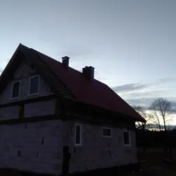 Niedokończony dom murowany z czerwonym dachem i dwoma kominami w szarym świetle zmierzchu, widoczne niezabezpieczone okna i brak elewacji.
