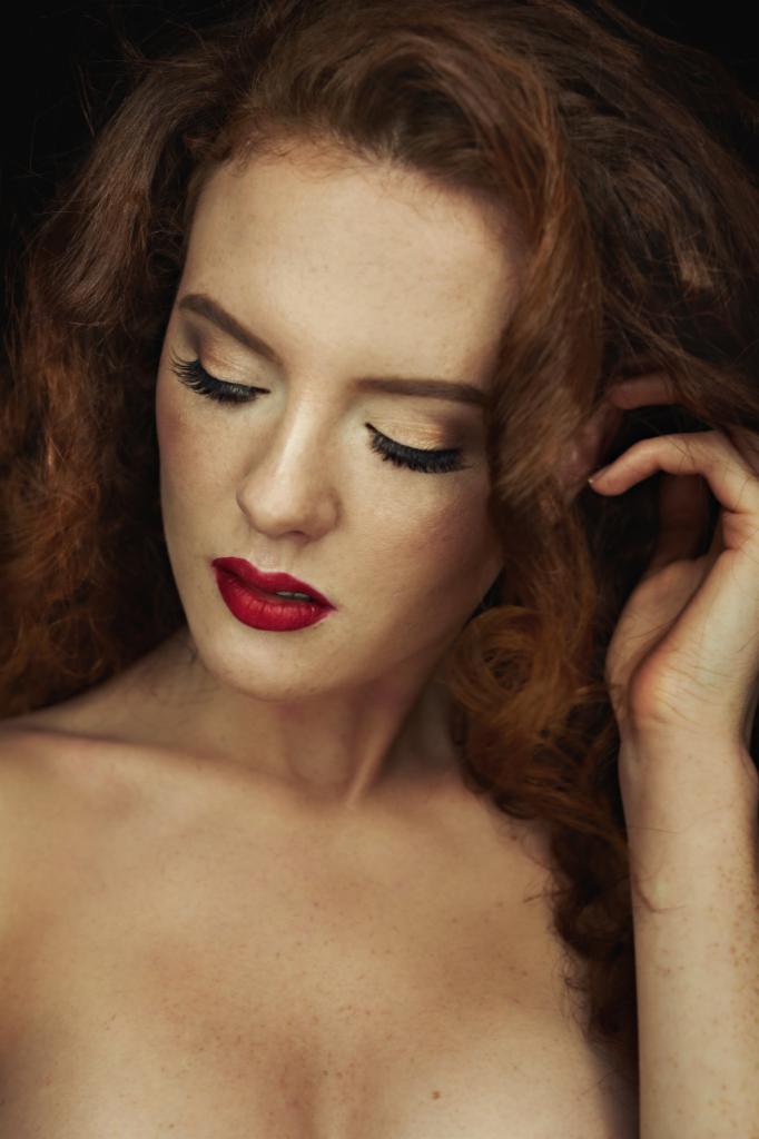 Portret kobiety o rudych włosach z intensywnym makijażem oczu i czerwonymi ustami, spoglądającej w dół, z dłonią we włosach.