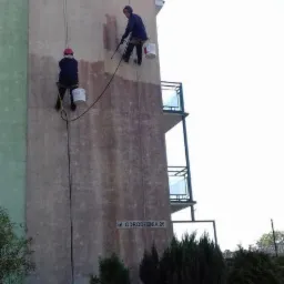 Dwóch alpinistów przemysłowych maluje elewację budynku przy użyciu lin i wiader z farbą, widoczny fragment pomalowanej ściany i tabliczka z adresem Ul. Odrodzenia 26.