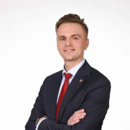 Portret uśmiechniętego mężczyzny w garniturze z czerwoną kravatą, stojącego na białym tle z założonymi rękami.