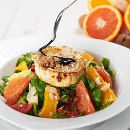 Restauracja Karpielówka - Sałatka z grillowanym serem kozim, pomarańczami, grejpfrutami i orzechami włoskimi, polana ciemnym sosem balsamicznym, serwowana na białym talerzu na tle białych desek.