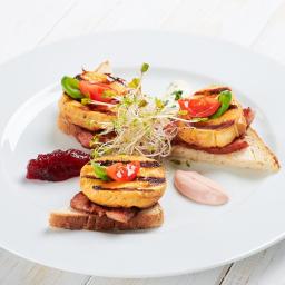 Restauracja Karpielówka - Elegancko podane kanapeczki na białym talerzu: grillowany ser halloumi na chlebie z wędliną, udekorowane pomidorkiem cherry, bazylią i kiełkami, z dodatkiem żurawiny i sosu.
