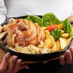 Restauracja Karpielówka - Pieczona golonka na kapuście z ziemniakami, pomidorem i sałatą, podana na drewnianej desce w żeliwnej patelni, trzymana przez osobę z widocznymi dłońmi z manicure.