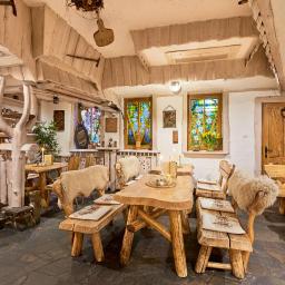 Restauracja Karpielówka - Rustykalne wnętrze restauracji z drewnianymi stołami i ławami obitymi owczą wełną, kolorowe witraże w oknach, kamienna podłoga i surowe drewniane wykończenie sufitu.