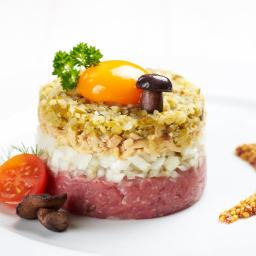 Restauracja Karpielówka - Elegancko ułożona warstwowa przystawka: tatar wołowy, cebula, tuńczyk, ogórek konserwowy, żółtko, grzybek, pomidorek koktajlowy i musztarda francuska na białym talerzu.