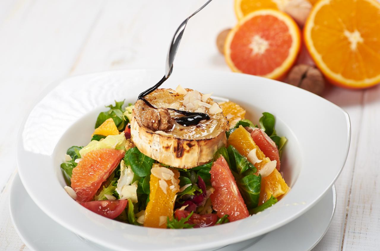 Sałatka z grillowanym serem kozim, pomarańczami, grejpfrutami i orzechami włoskimi, polana ciemnym sosem balsamicznym, serwowana na białym talerzu na tle białych desek.