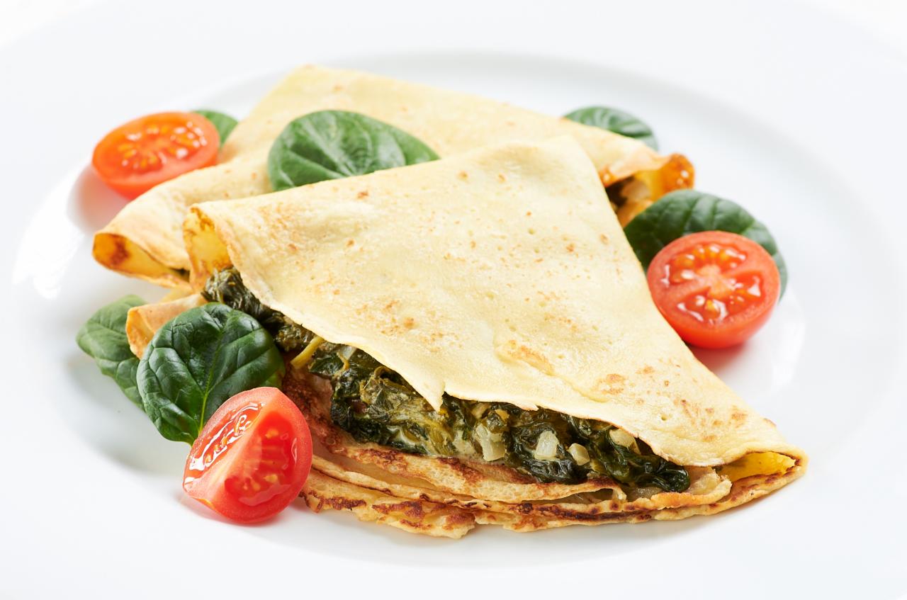 Delikatne naleśniki wypełnione szpinakiem i cebulą, udekorowane świeżymi liśćmi szpinaku i połówkami pomidorków koktajlowych na białym talerzu.