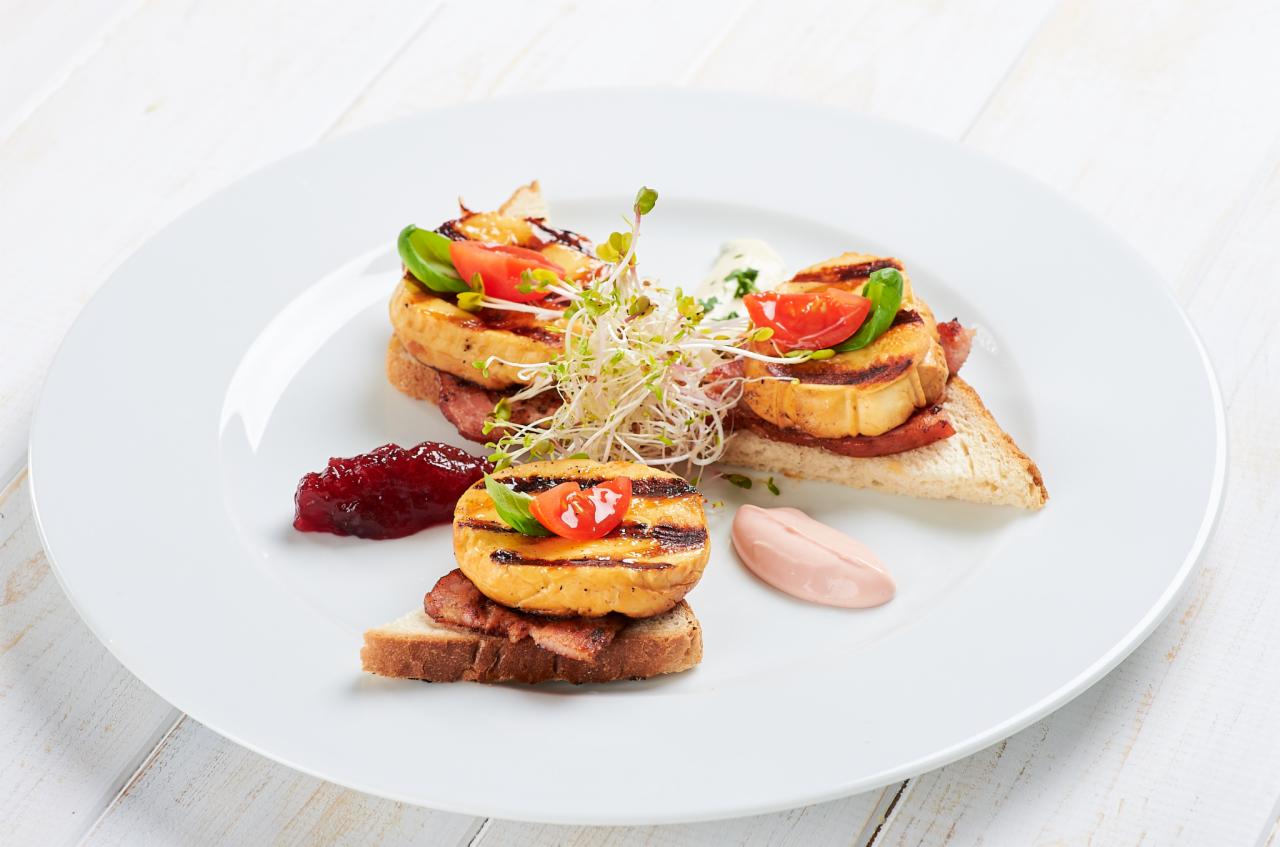 Elegancko podane kanapeczki na białym talerzu: grillowany ser halloumi na chlebie z wędliną, udekorowane pomidorkiem cherry, bazylią i kiełkami, z dodatkiem żurawiny i sosu.