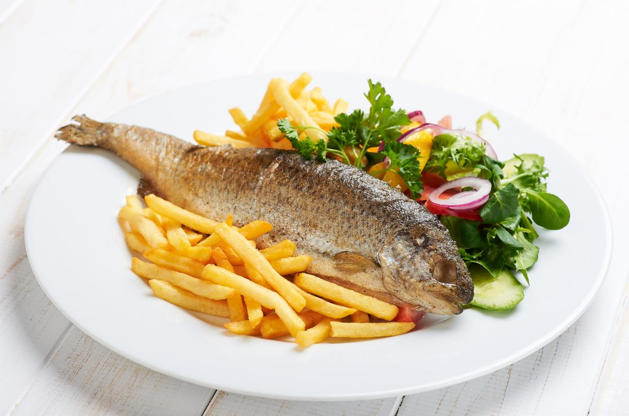 Upieczona pstrąg serwowany na białym talerzu z frytkami i sałatką z pomarańczy, czerwonej cebuli i zielonych liści, ułożony na białym drewnianym stole.