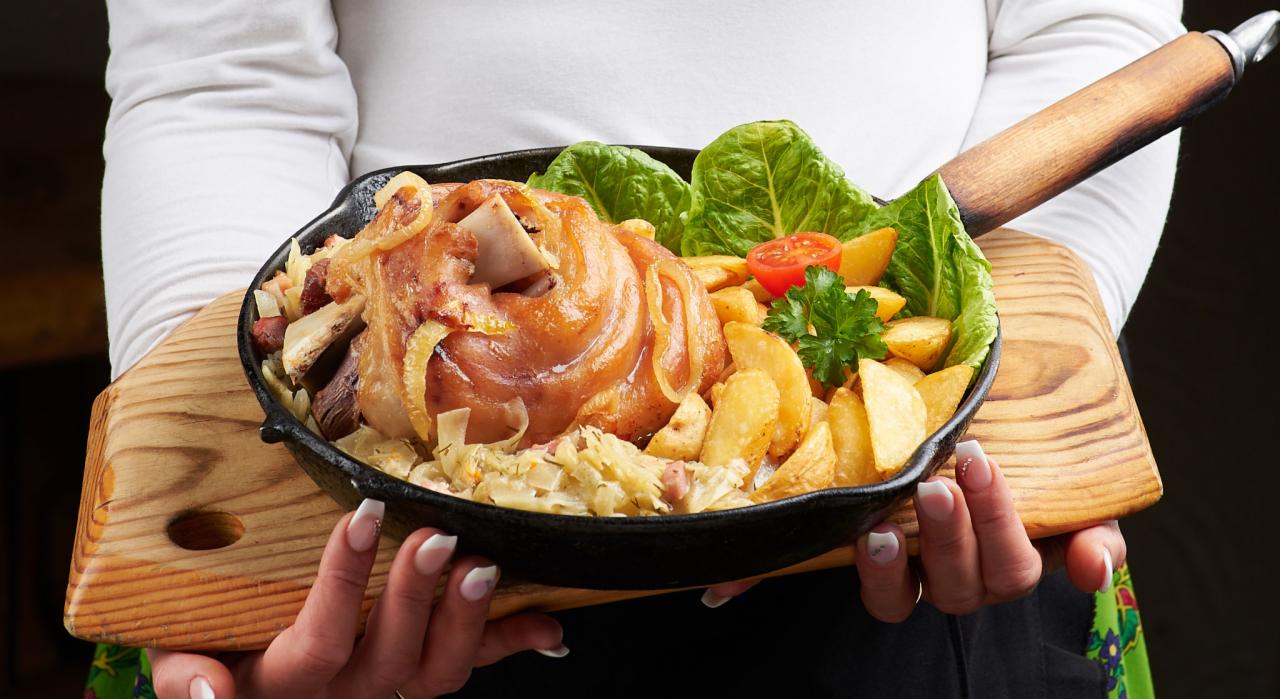 Pieczona golonka na kapuście z ziemniakami, pomidorem i sałatą, podana na drewnianej desce w żeliwnej patelni, trzymana przez osobę z widocznymi dłońmi z manicure.