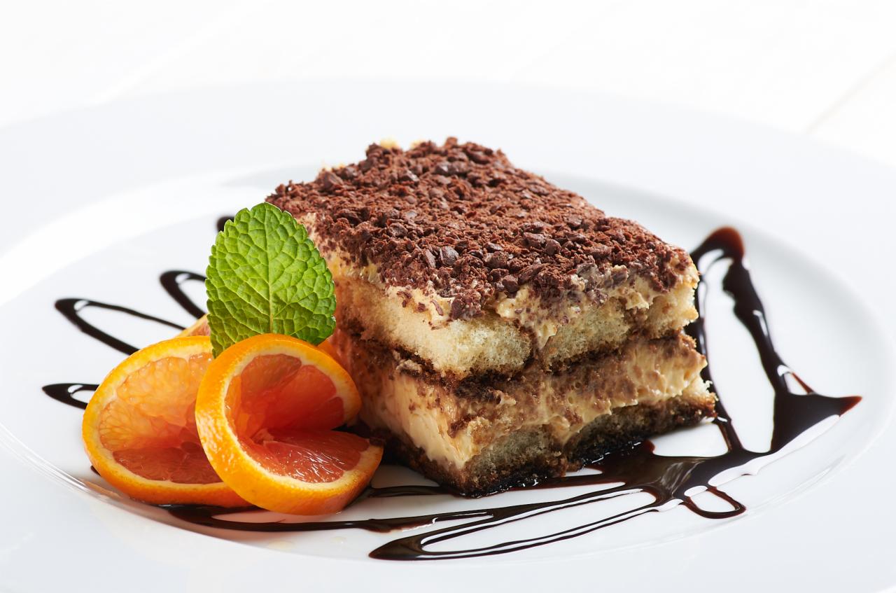 Elegancko podany deser tiramisu z posypką czekoladową, dekorowany listkiem mięty i dwoma półplasterkami pomarańczy, polany sosem czekoladowym na białym talerzu.