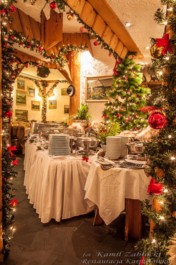 Świątecznie udekorowany stół bufetowy z podgrzewaczami i stosami talerzy w rustykalnej restauracji, ozdobiony choinkami i lampkami.