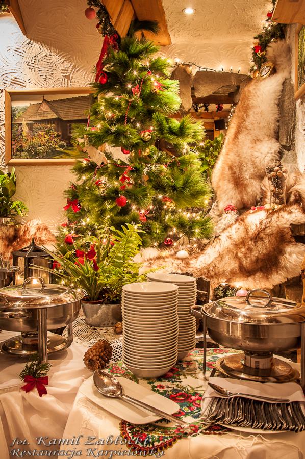 Uroczysty stół szwedzki z dekoracjami świątecznymi, w tle choinka i futrzana dekoracja, zastawa stołowa i podgrzewacze potraw.