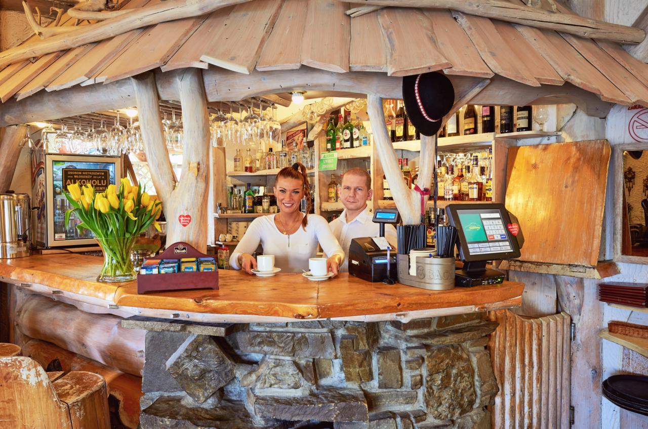 Uśmiechnięta barmanka i barman za rustykalnym barem z drewna i kamienia, serwujący kawę, widoczne butelki alkoholi i góralski kapelusz.