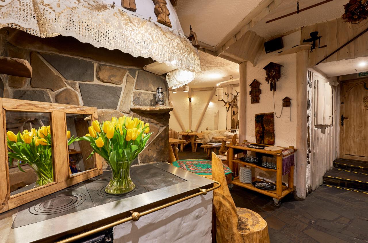 Rustykalne wnętrze restauracji z kamienną ścianą, piecem kaflowym zwieńczonym wazonem z żółtymi tulipanami, drewnianymi meblami i dekoracjami, tworzące ciepłą, tradycyjną atmosferę.