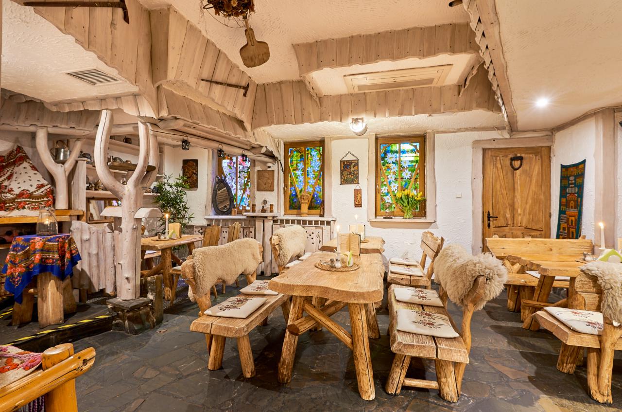 Rustykalne wnętrze restauracji z drewnianymi stołami i ławami obitymi owczą wełną, kolorowe witraże w oknach, kamienna podłoga i surowe drewniane wykończenie sufitu.