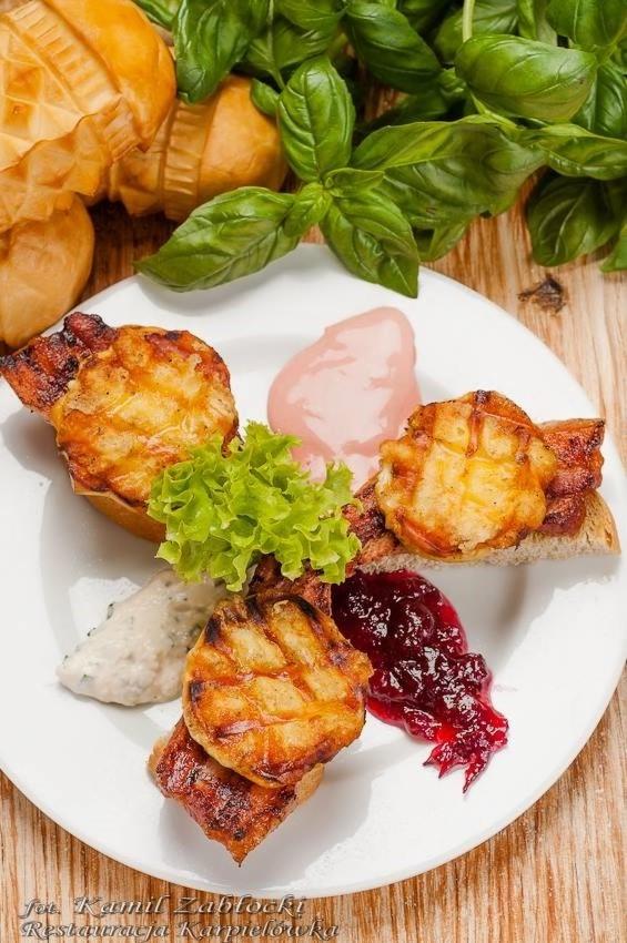 Grillowany oscypek na grzance z żurawiną, sosem tatarskim i różowym sosem, udekorowany sałatą i bazylią, podany na białym talerzu na drewnianym stole.