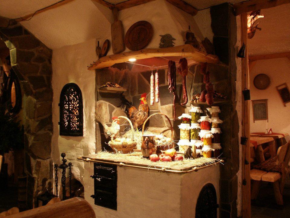 Wnętrze restauracji w rustykalnym stylu z piecem, na którym znajdują się słoiki z przetworami, wędliny, koszyki z jajkami i jabłka, a także dekoracyjne figurki kogutów.