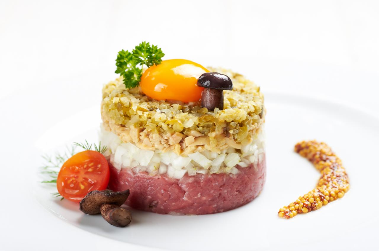 Elegancko ułożona warstwowa przystawka: tatar wołowy, cebula, tuńczyk, ogórek konserwowy, żółtko, grzybek, pomidorek koktajlowy i musztarda francuska na białym talerzu.