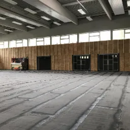 Wnętrze hali sportowej w trakcie remontu: widoczna konstrukcja ścian z płyt OSB na stelażu, podłoga z widocznymi śladami po kleju, podnośnik koszowy w rogu sali i oświetlenie sufitowe