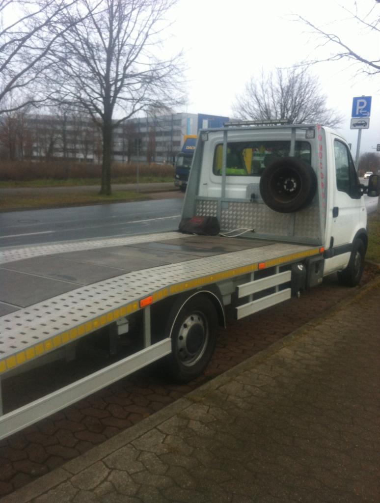 Biała laweta z platformą najazdową zaparkowana przy ulicy w pochmurny dzień. Z tyłu, na metalowej konstrukcji, zamontowane koło zapasowe. Widoczny znak parkingu dla lawet.