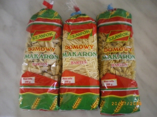 Trzy przezroczyste opakowania makaronu 'Domowy Makaron Bartos' o różnej gramaturze i kształtach: muszelki, nitki i świderki, ustawione obok siebie na jasnym tle.