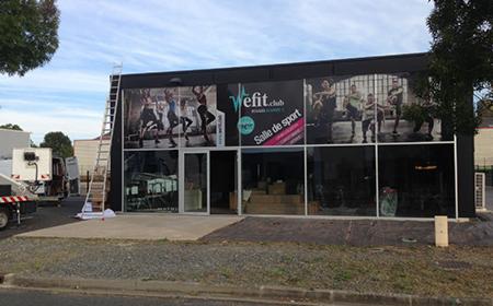 Fasada budynku z dużymi oknami ozdobiona grafiką reklamową siłowni 'Vefit.club', widoczna drabina i samochód dostawczy sugerujące montaż lub demontaż, pochmurne niebo w tle.