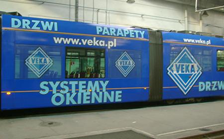 Niebieski tramwaj z reklamą firmy Veka na drzwiach, parapetach i systemach okiennych, widoczne logo firmy w białej ramce.