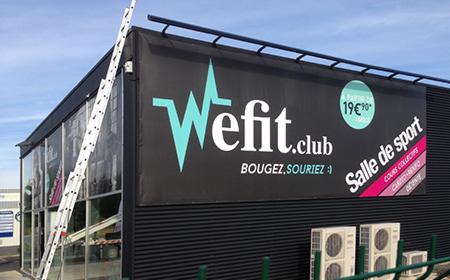 Czarny budynek z dużym banerem reklamowym 'Wefit.club' z turkusowym logo w kształcie pulsu, hasłem 'BOUGEZ, SOURIEZ :)' oraz informacją o cenie. Przy budynku oparta drabina, widoczne klimatyzatory.