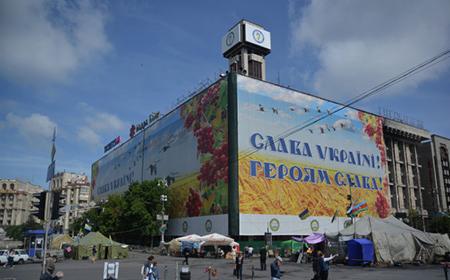 Wielkoformatowy baner z ukraińskim hasłem 'Sława Ukraini! Herojam Sława!' na budynku z wieżą zegarową, widok z placu z rozstawionymi namiotami.