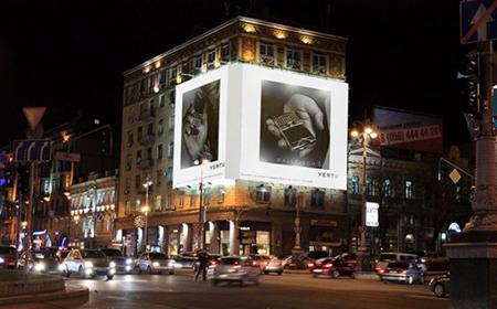 Nocny widok na budynek z podświetlonym billboardem na dachu, prezentującym czarno-białe zdjęcia produktu marki Vertu, z ruchem ulicznym w tle.