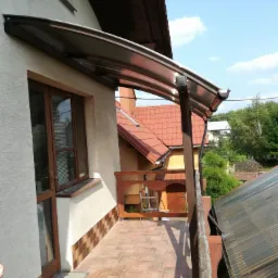 Zadaszony balkon z brązową balustradą, brązowymi płytkami na podłodze i przezroczystym zadaszeniem nad drzwiami balkonowymi, widok na sąsiedni dom z czerwonym dachem.