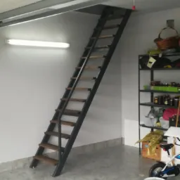 Stalowa drabina strychowa z drewnianymi stopniami, zamontowana w garażu, widok na otwarty właz na strych i półki z zapasami w tle.