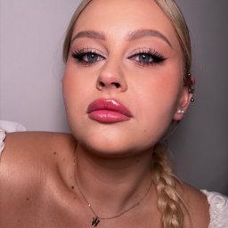 Jagoda Słomka INSTRUKTOR RZĘS - Wizaż, Stylizacja brwi i rzęs - Portret kobiety z profesjonalnym makijażem: wyraziste, czarne kreski na powiekach, podkreślone różem policzki, błyszczące, różowe usta i blond warkocz. Widoczne kolczyki w uchu i złoty łańcuszek...