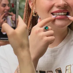 Uśmiechnięta kobieta z ozdobami na zębach, prezentująca efekt stylizacji, fotografowana telefonem; widoczne dłonie z ozdobnym pierścieniem i manicure.