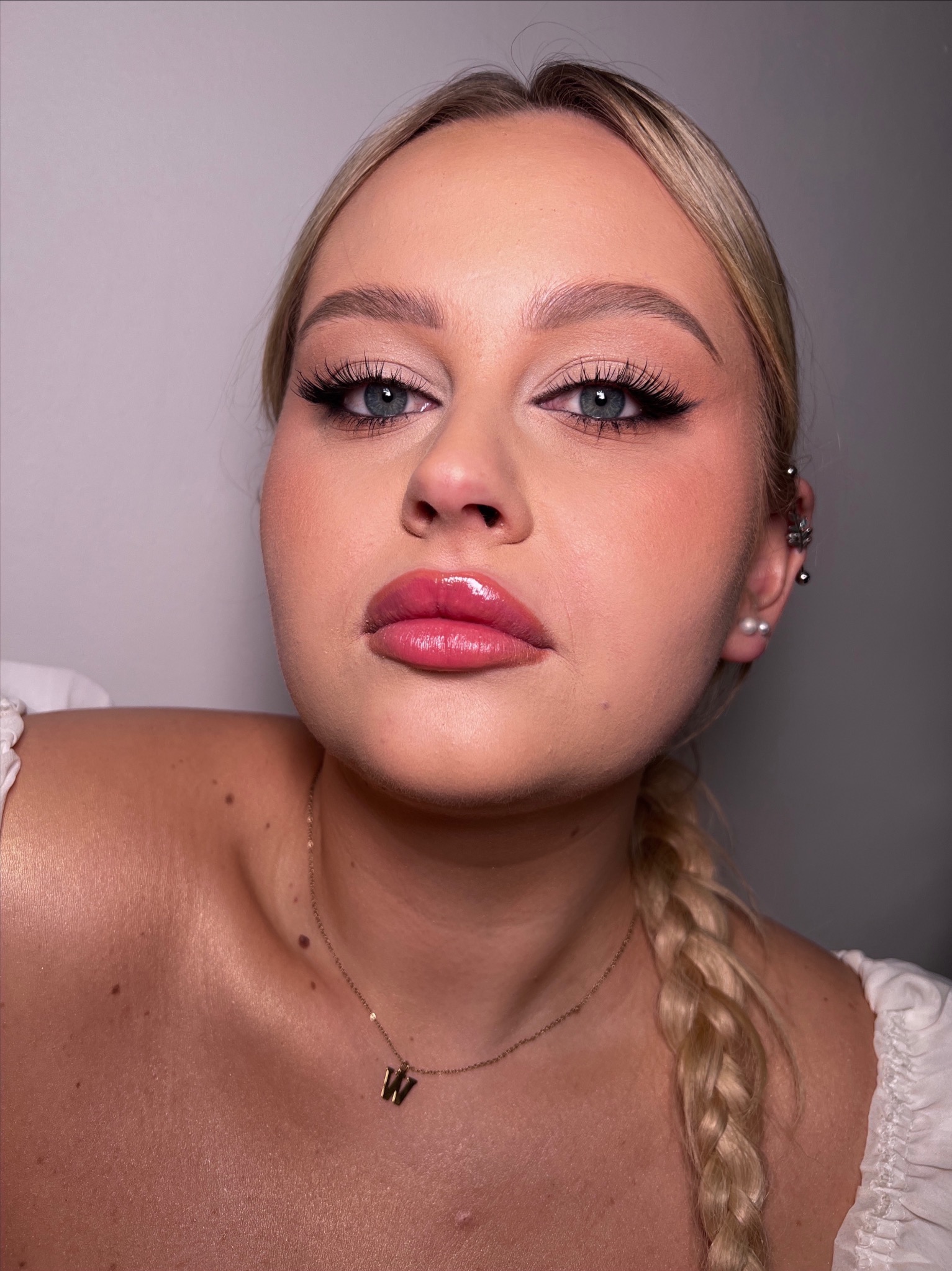 Portret kobiety z profesjonalnym makijażem: wyraziste, czarne kreski na powiekach, podkreślone różem policzki, błyszczące, różowe usta i blond warkocz. Widoczne kolczyki w uchu i złoty łańcuszek...